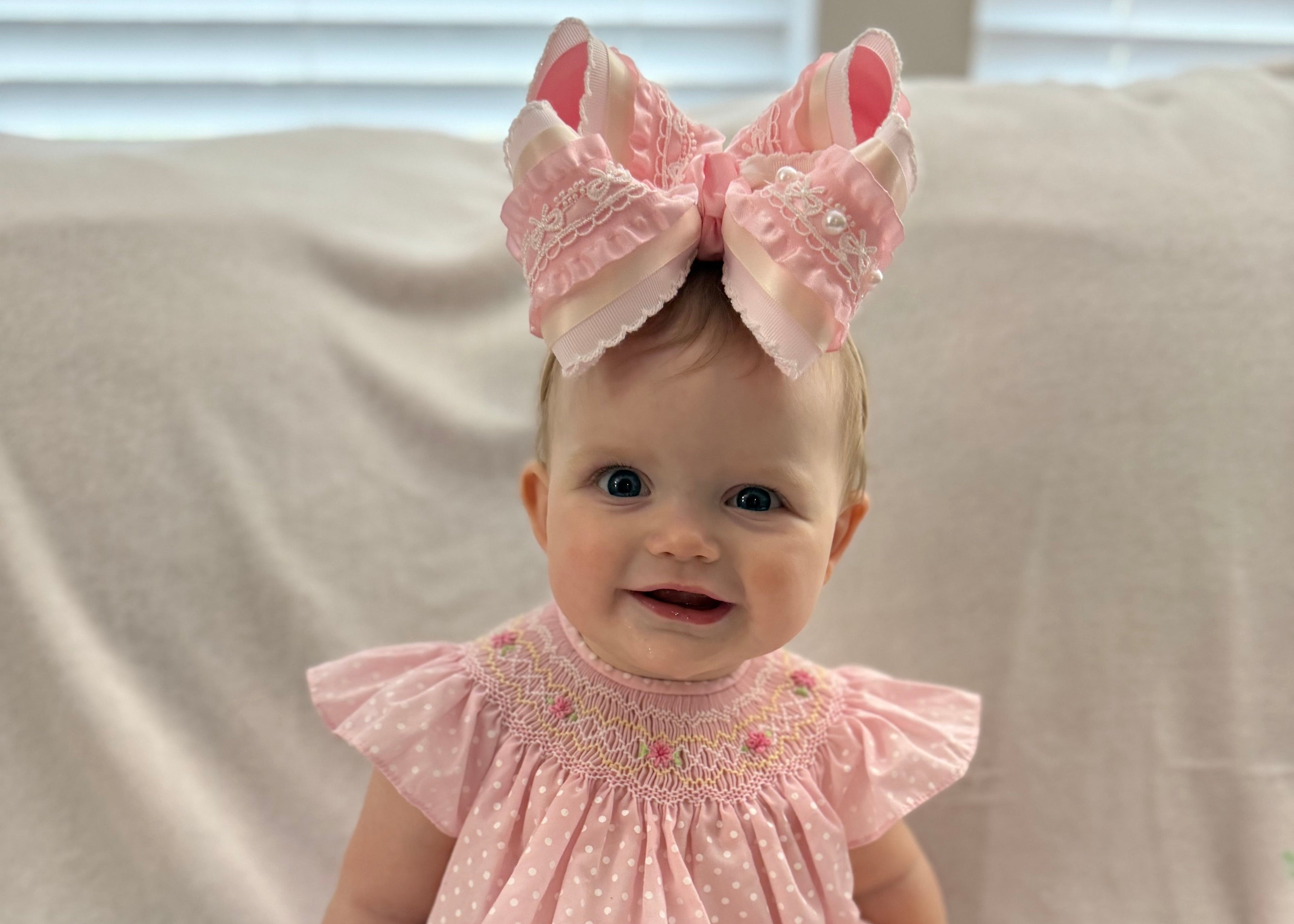 Baby wearing a pink dress and large bow sitting on a light-colored couch.